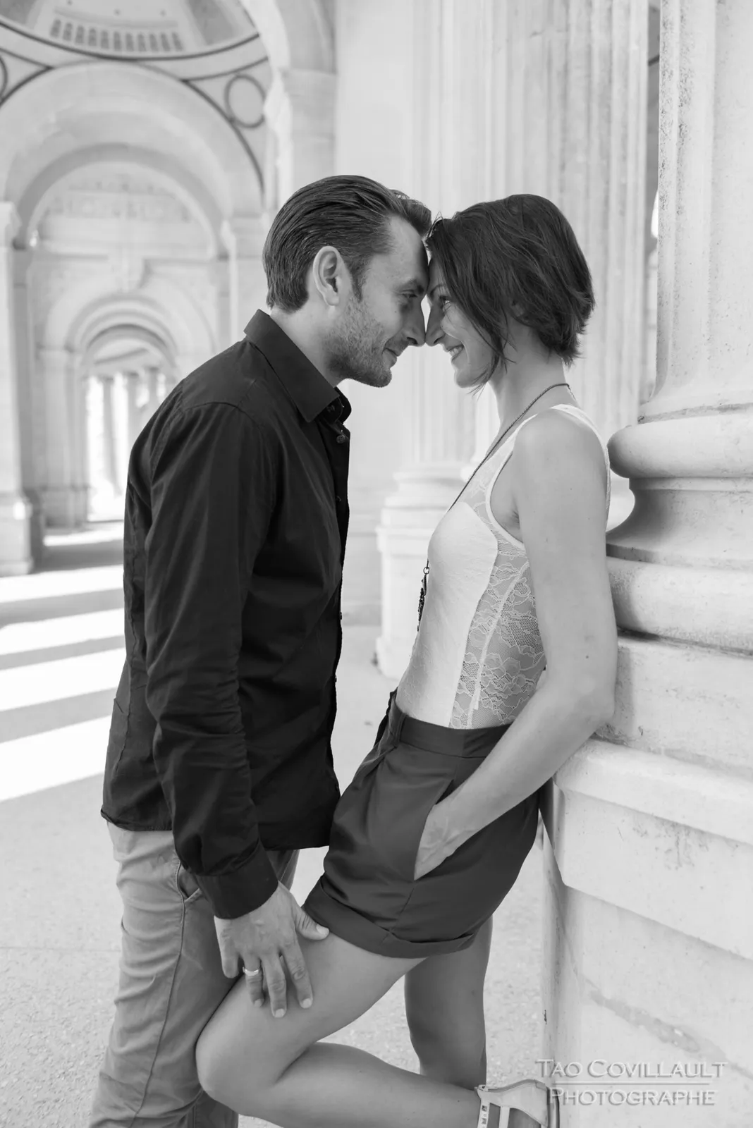 Black-and-white photograph: couple embracing and posing among stone columns.