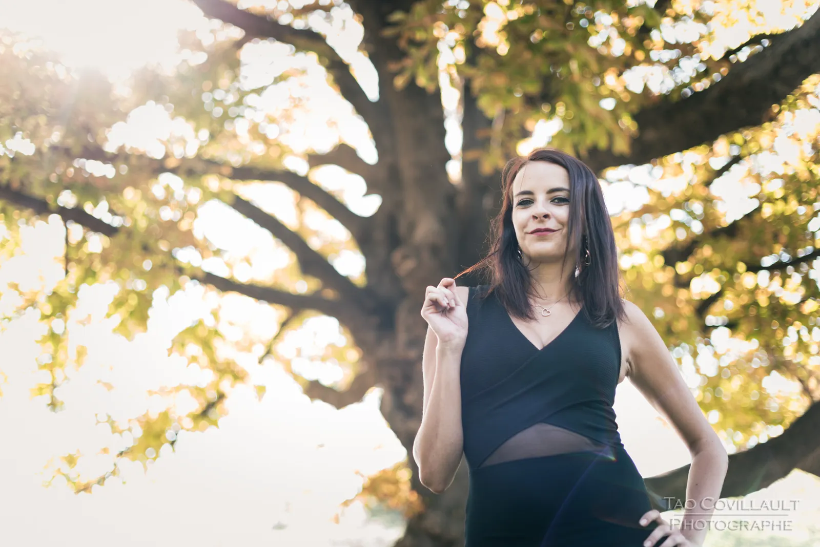 Woman in a black dress standing beneath a leafy tree.