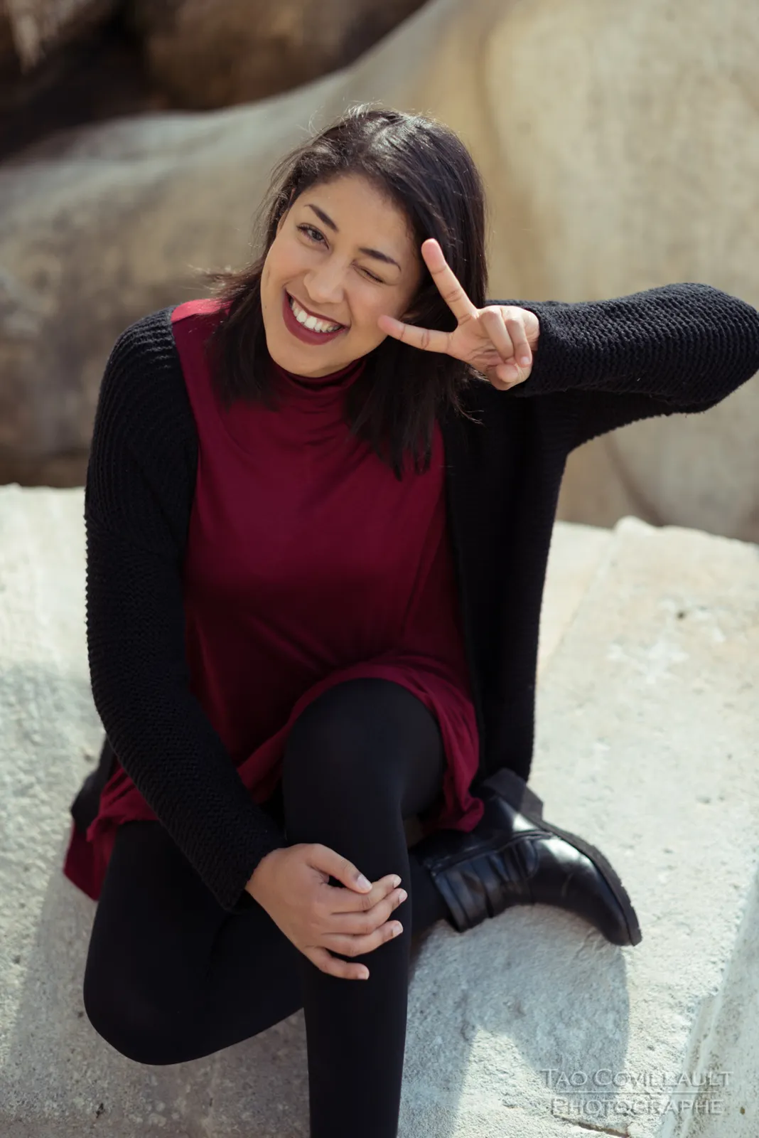 Woman in a burgundy dress and dark coat seated beside a carved stone sculpture.