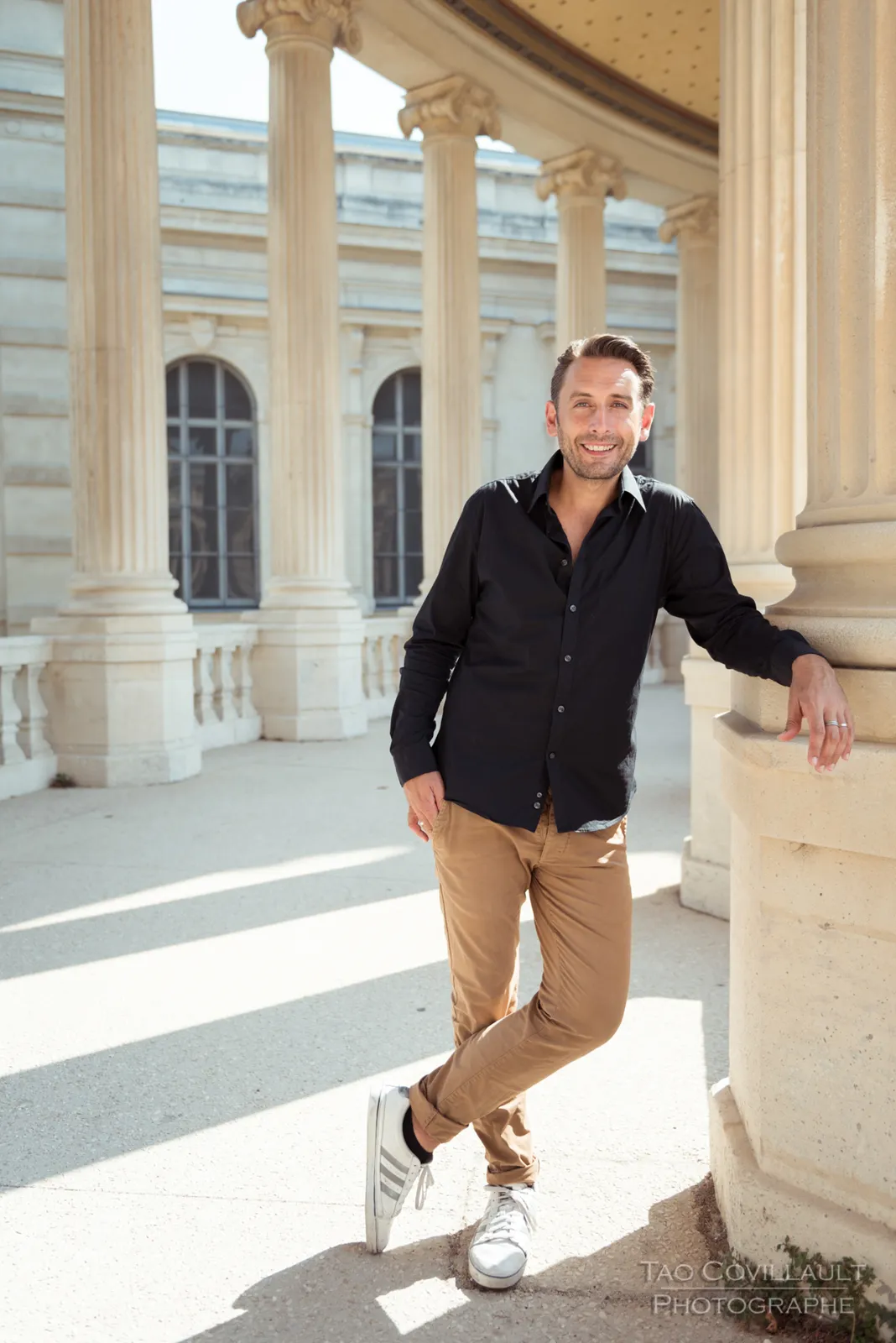 Man in a dark shirt and tan trousers posing in a colonnade.