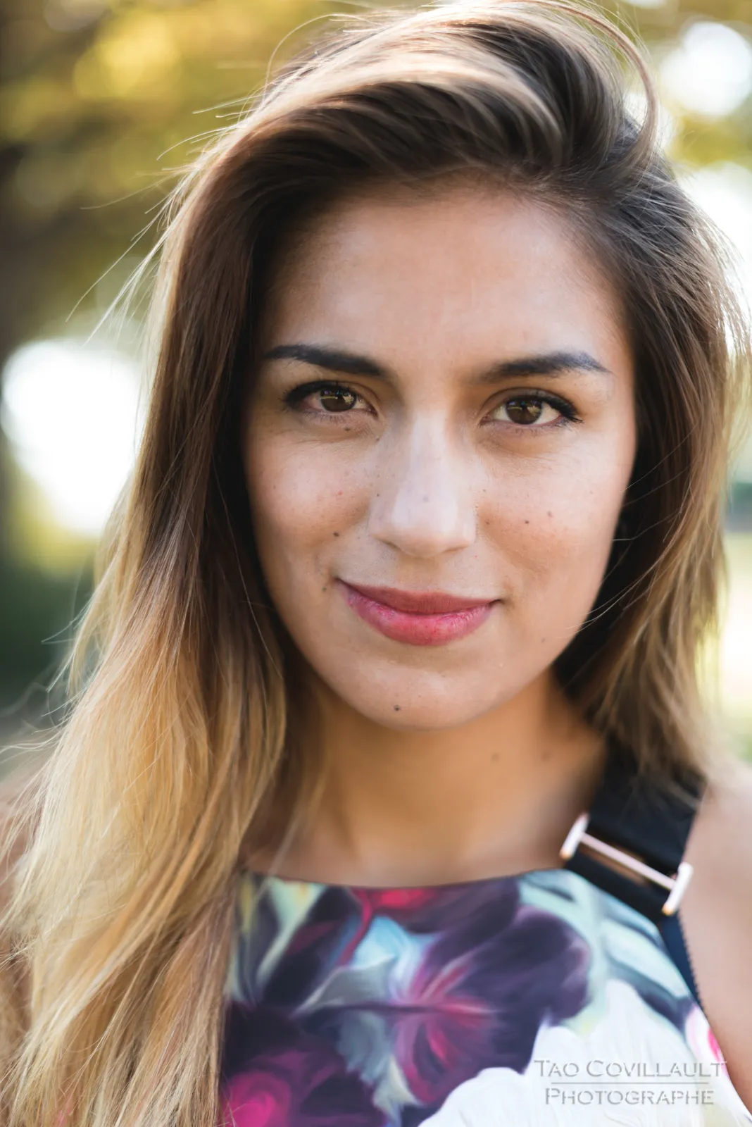 Woman in a floral dress posing beneath leafy trees.