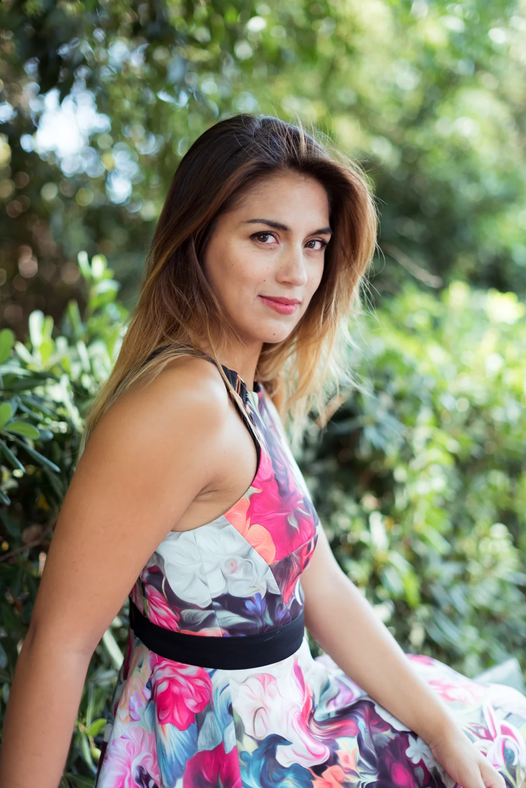 Woman in a floral dress posing beside a green park bench.