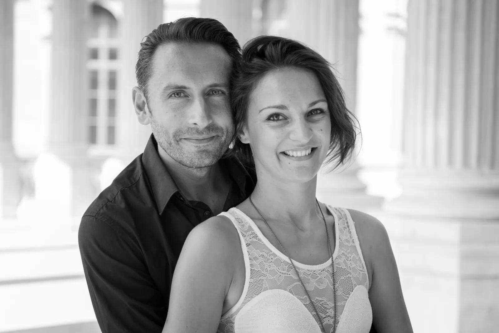 Black-and-white photograph: couple embracing and posing among stone columns.