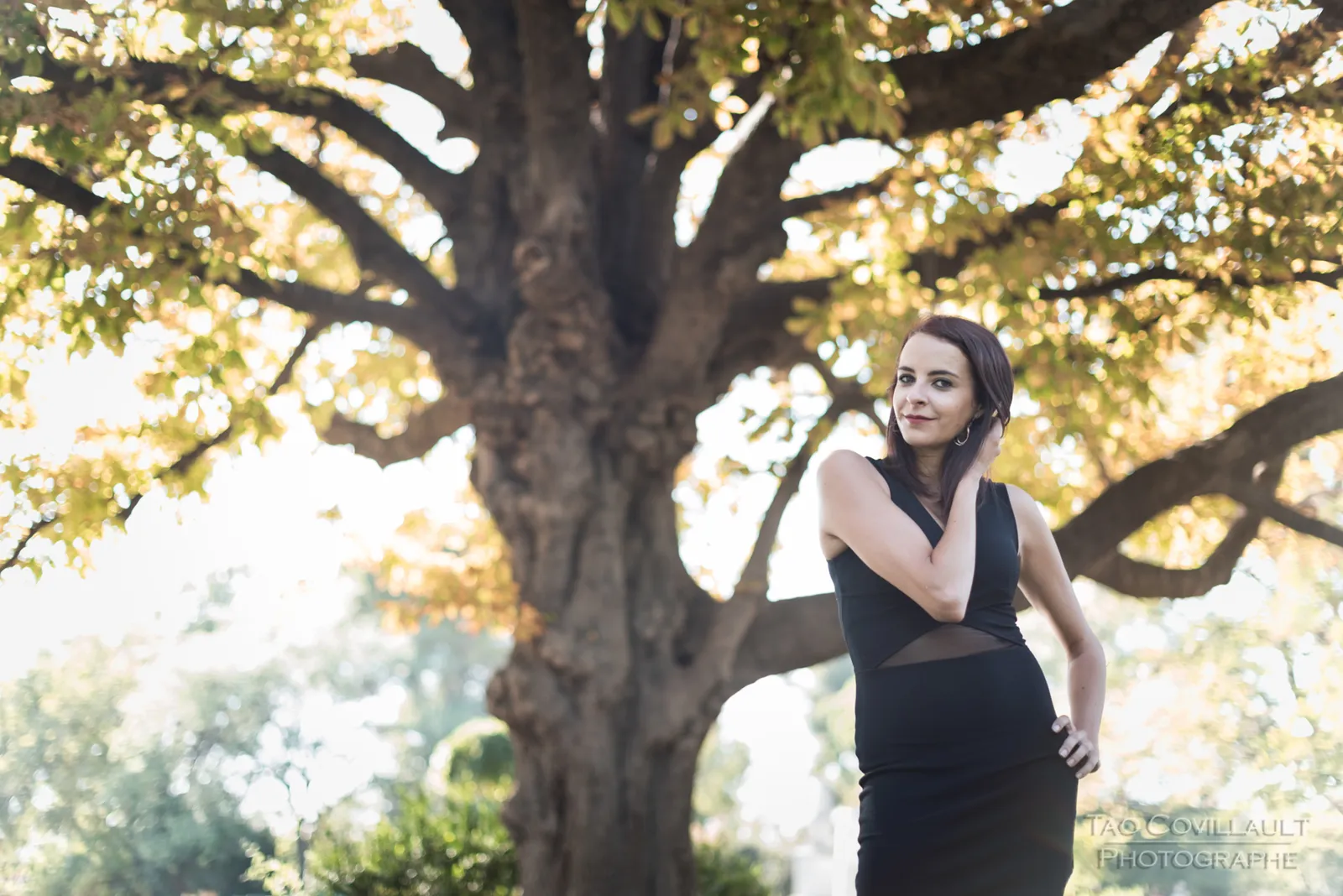 Woman in a black dress standing beneath a leafy tree.