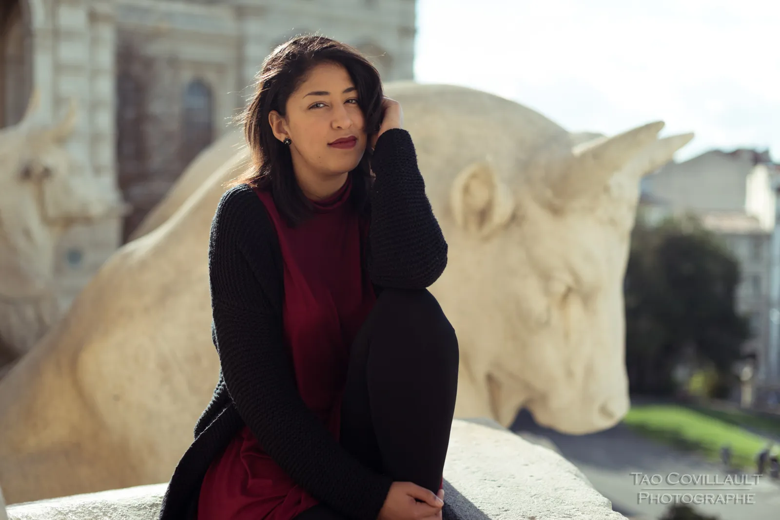Woman in a burgundy dress and dark coat seated beside a carved stone sculpture.