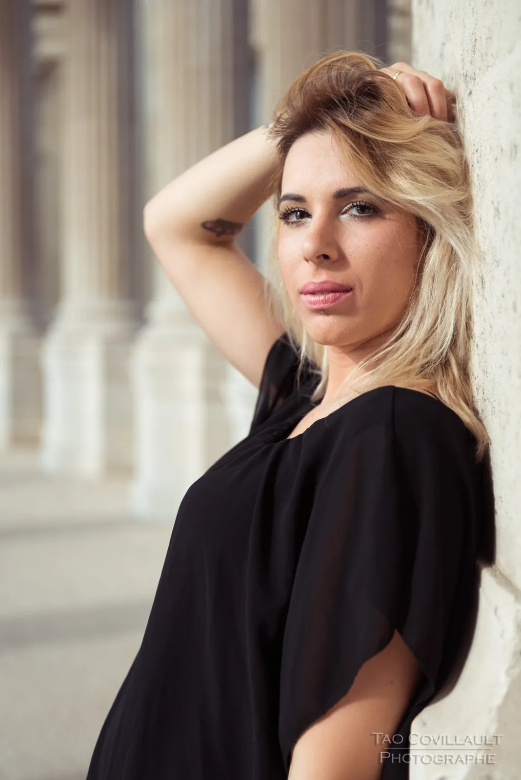 Woman in a black top and jeans posing along a stone colonnade.