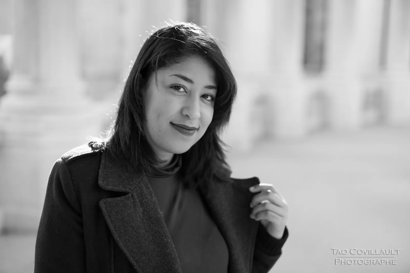 Black-and-white photograph: woman in a dark coat and burgundy dress posing among pale stone columns.