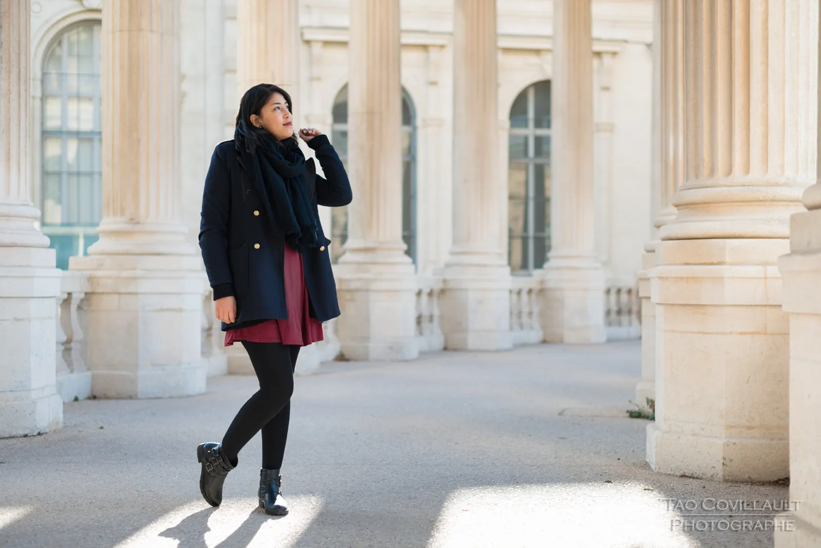 Woman in a dark coat and burgundy dress posing among pale stone columns.
