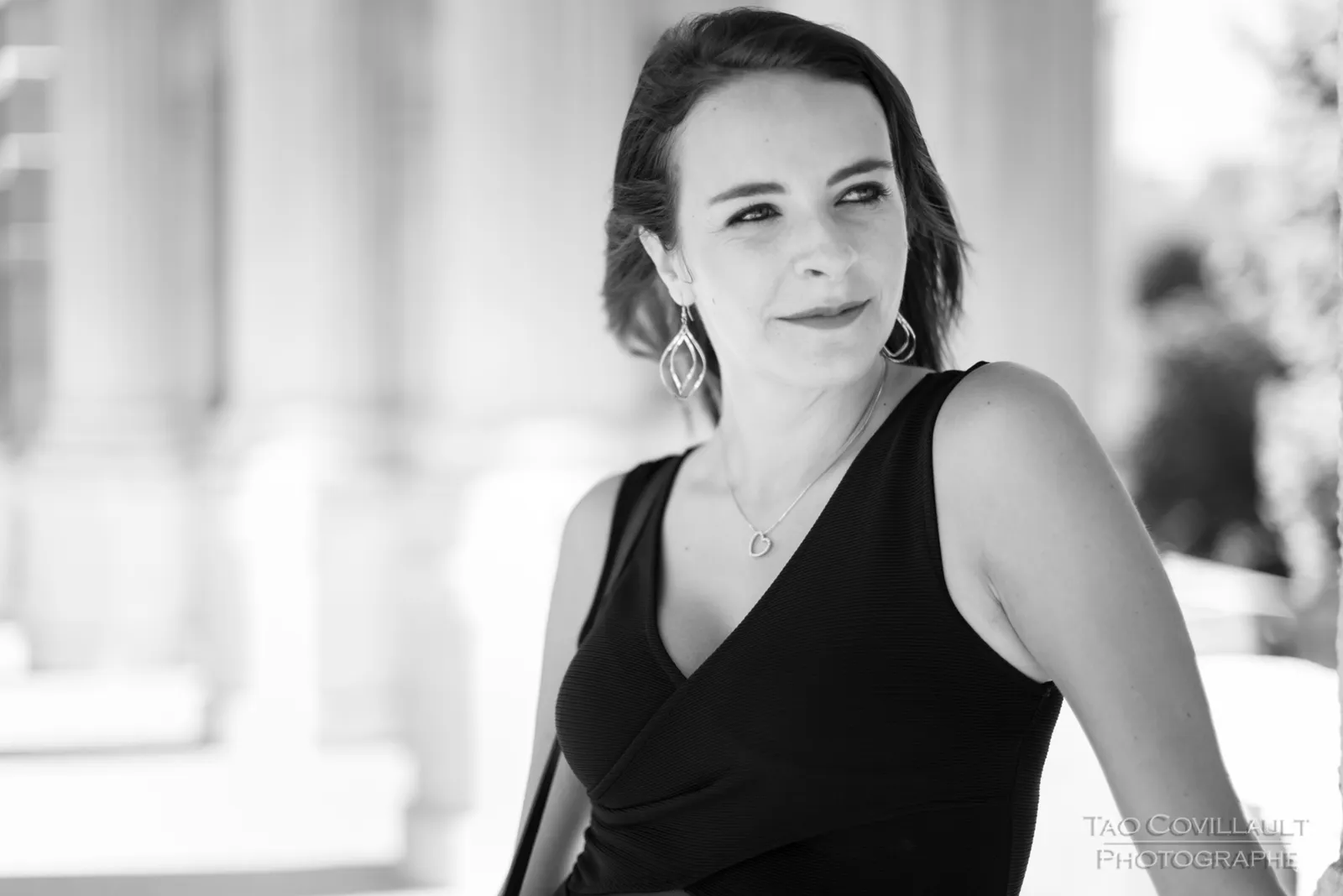 Black-and-white photograph: woman in a black dress posing along a sunlit colonnade.