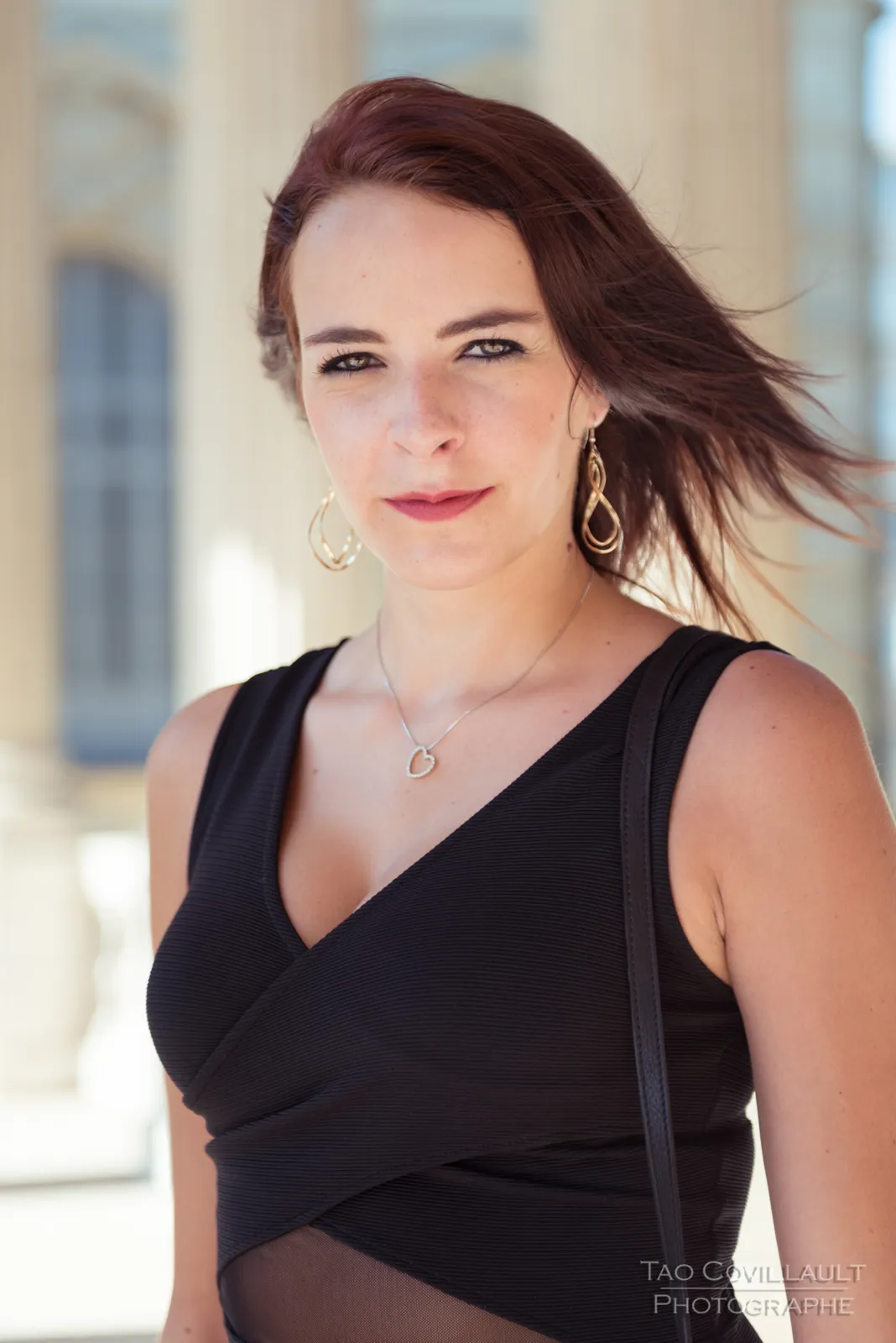 Woman in a black dress posing along a sunlit colonnade.