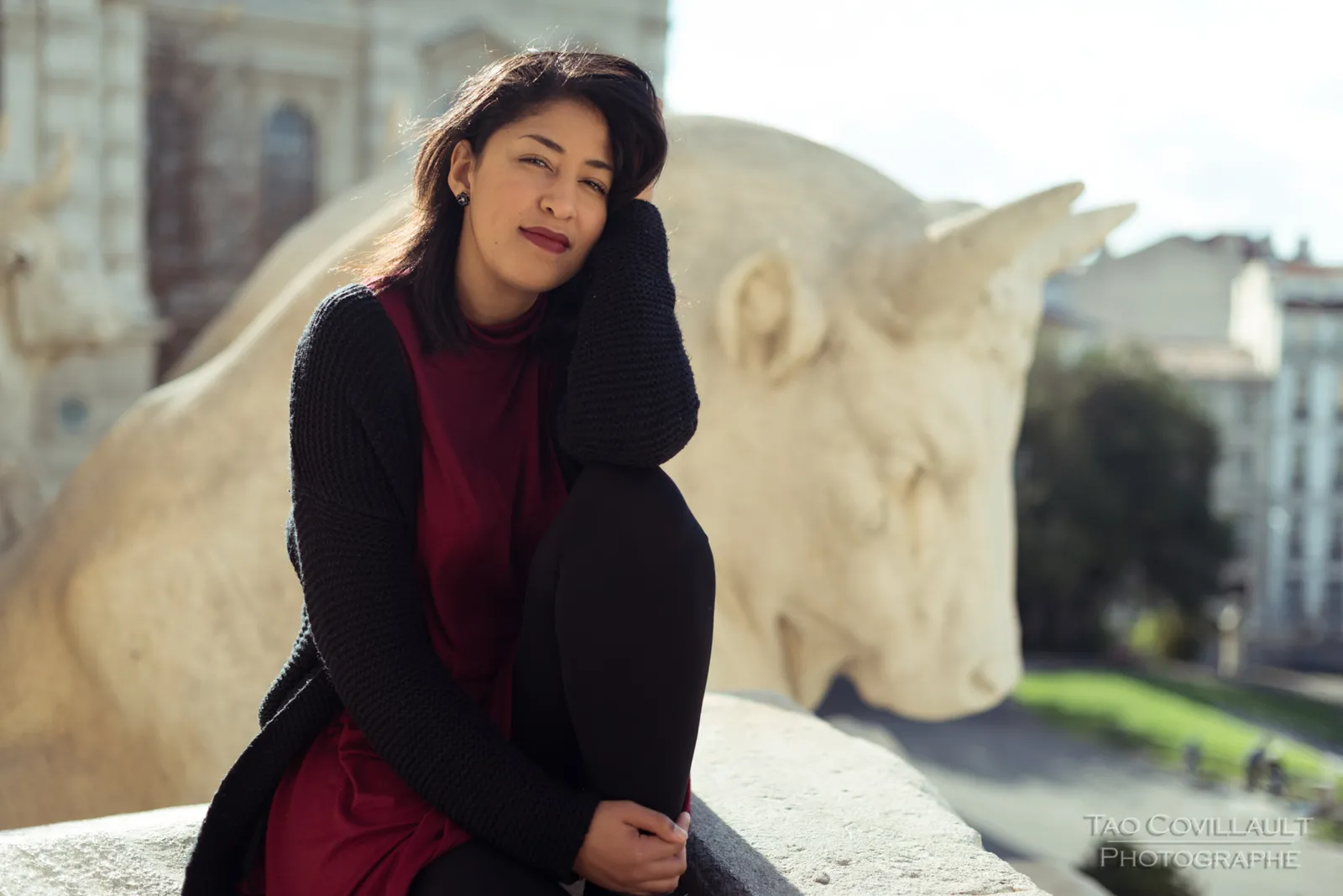 Woman in a burgundy dress and dark coat seated beside a carved stone sculpture.
