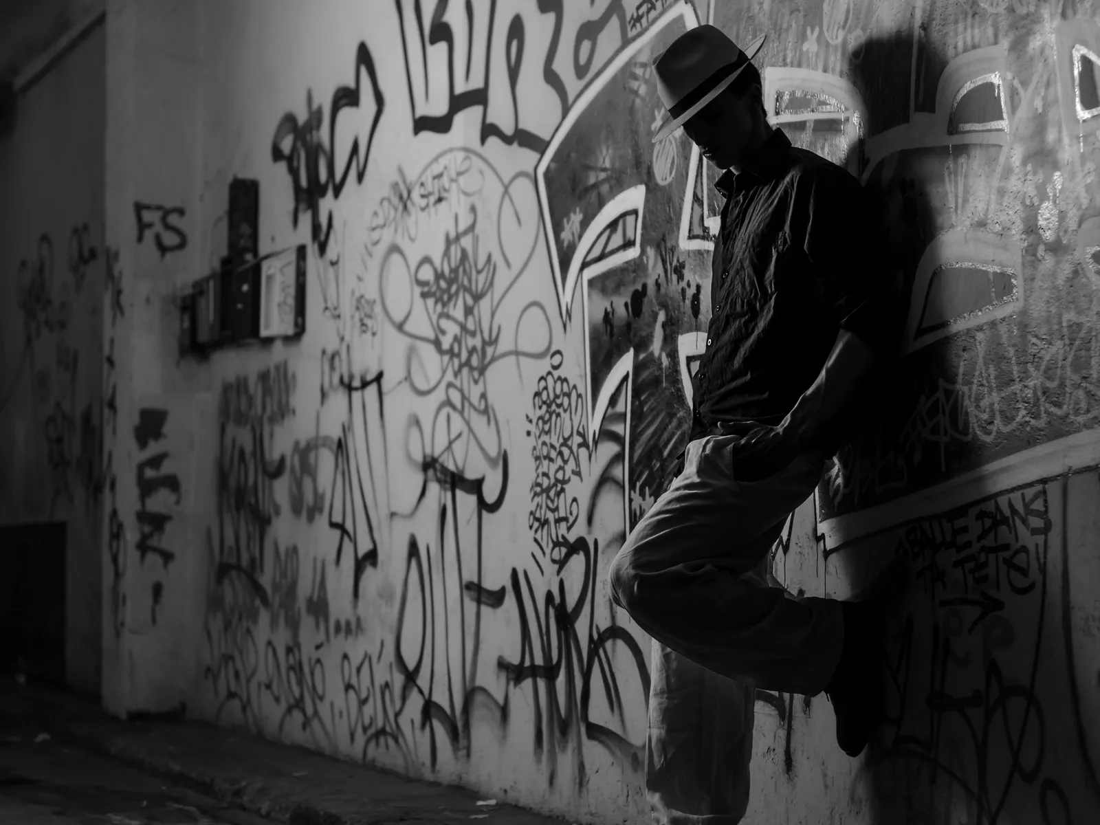 Shadowed person in a hat leaning against a graffiti-covered wall