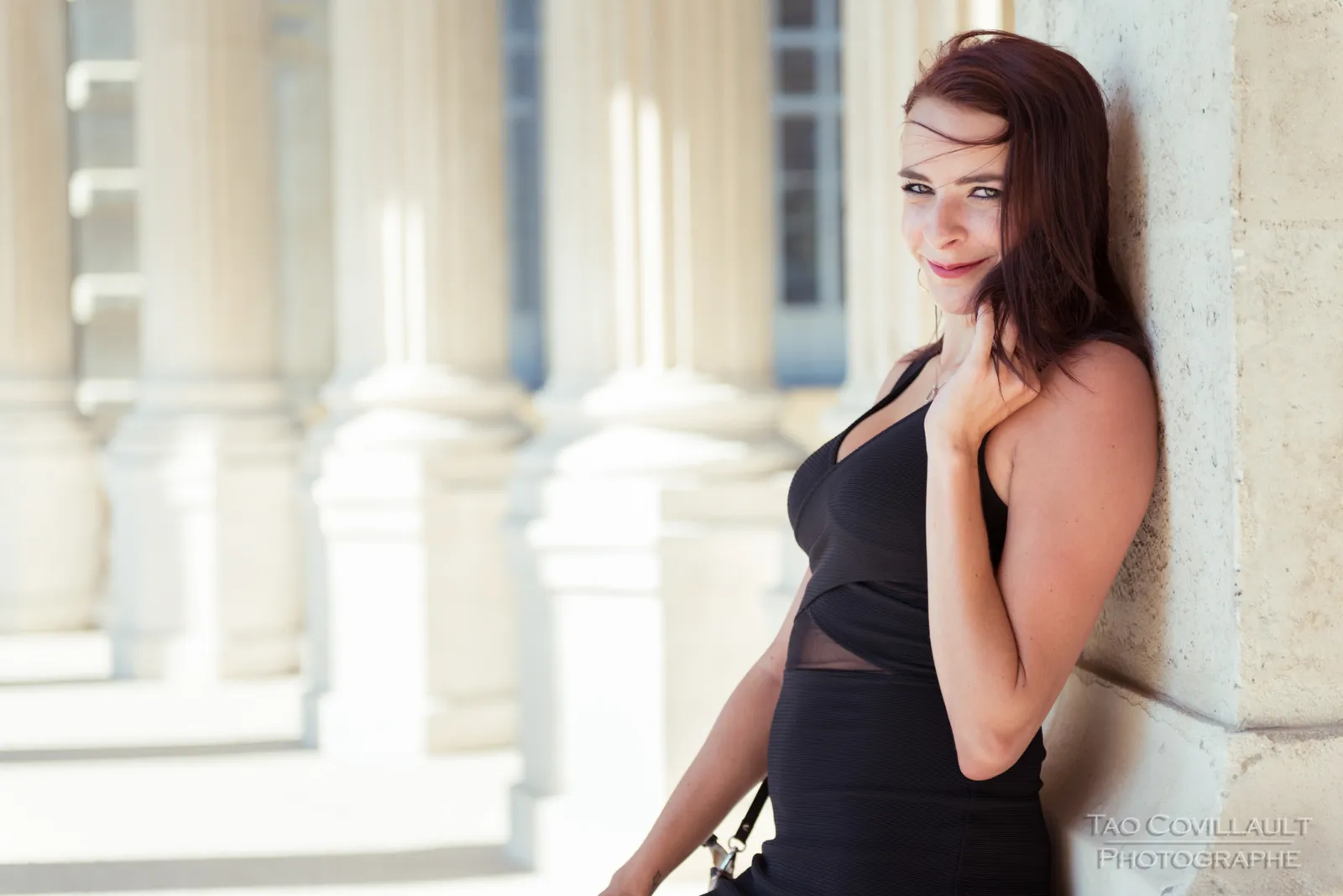 Woman in a black dress posing along a sunlit colonnade.