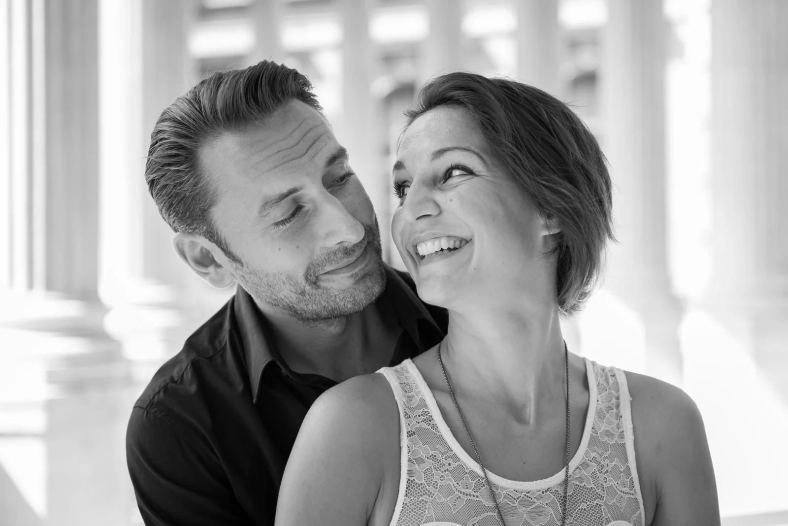 Black-and-white photograph: couple embracing and posing among stone columns.