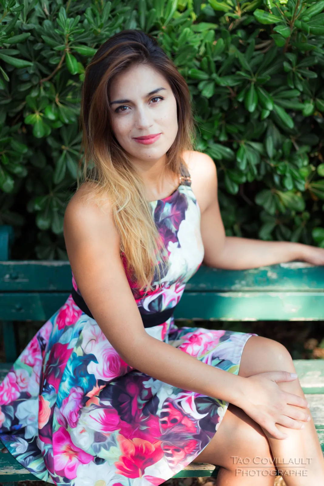 Woman in a floral dress posing beside a green park bench.