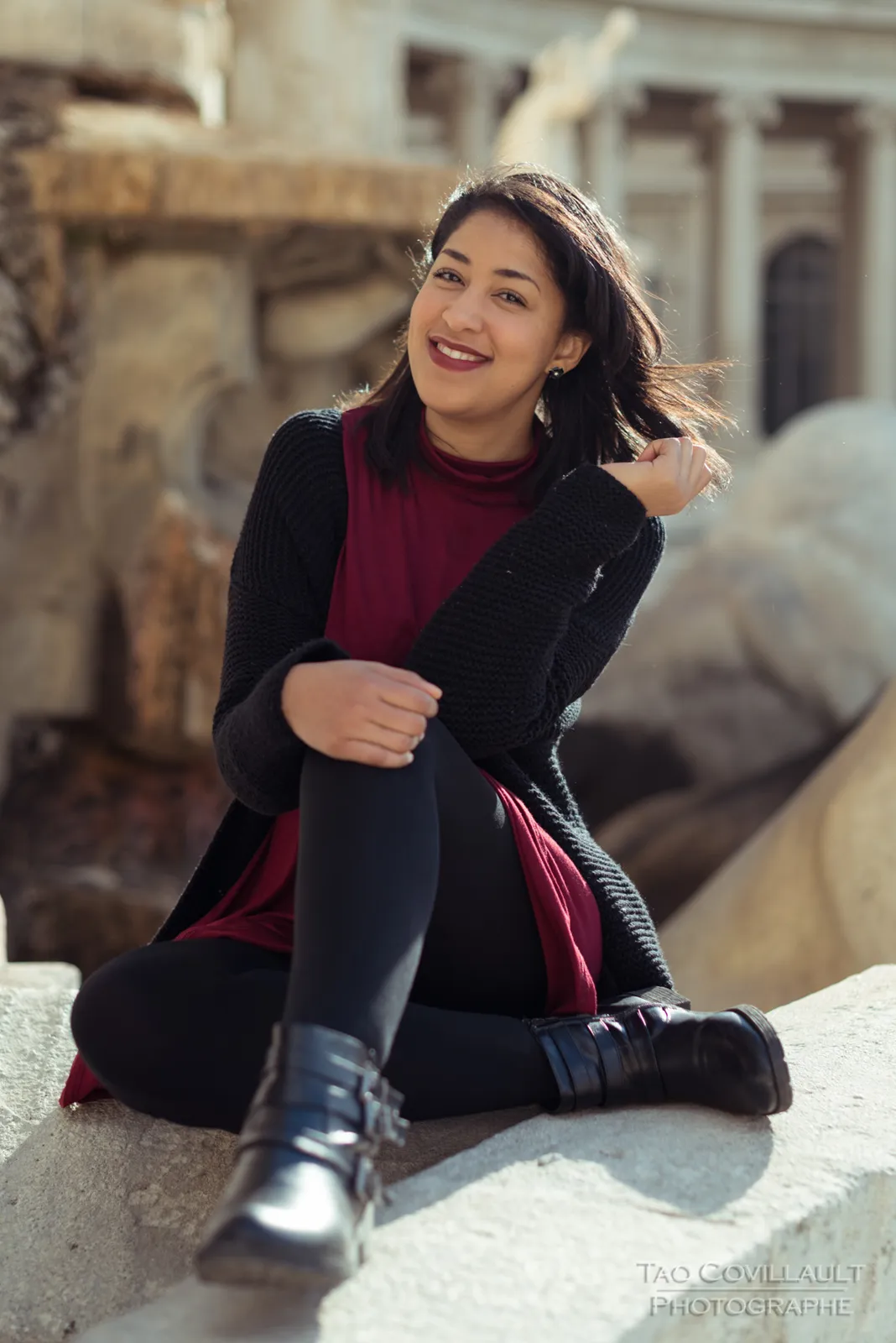 Woman in a burgundy dress and dark coat seated beside a carved stone sculpture.