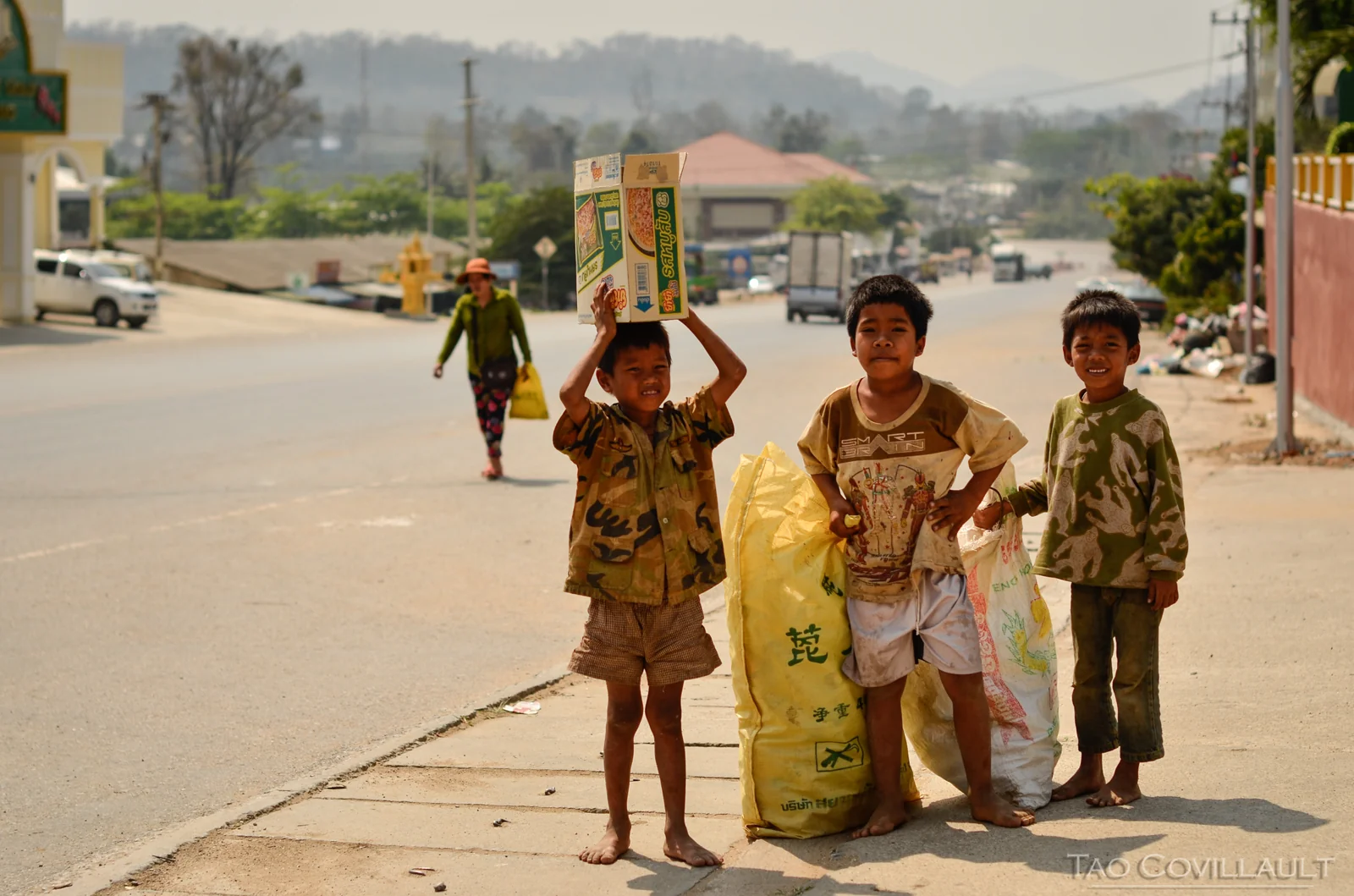 Three children carrying items beside a paved road.