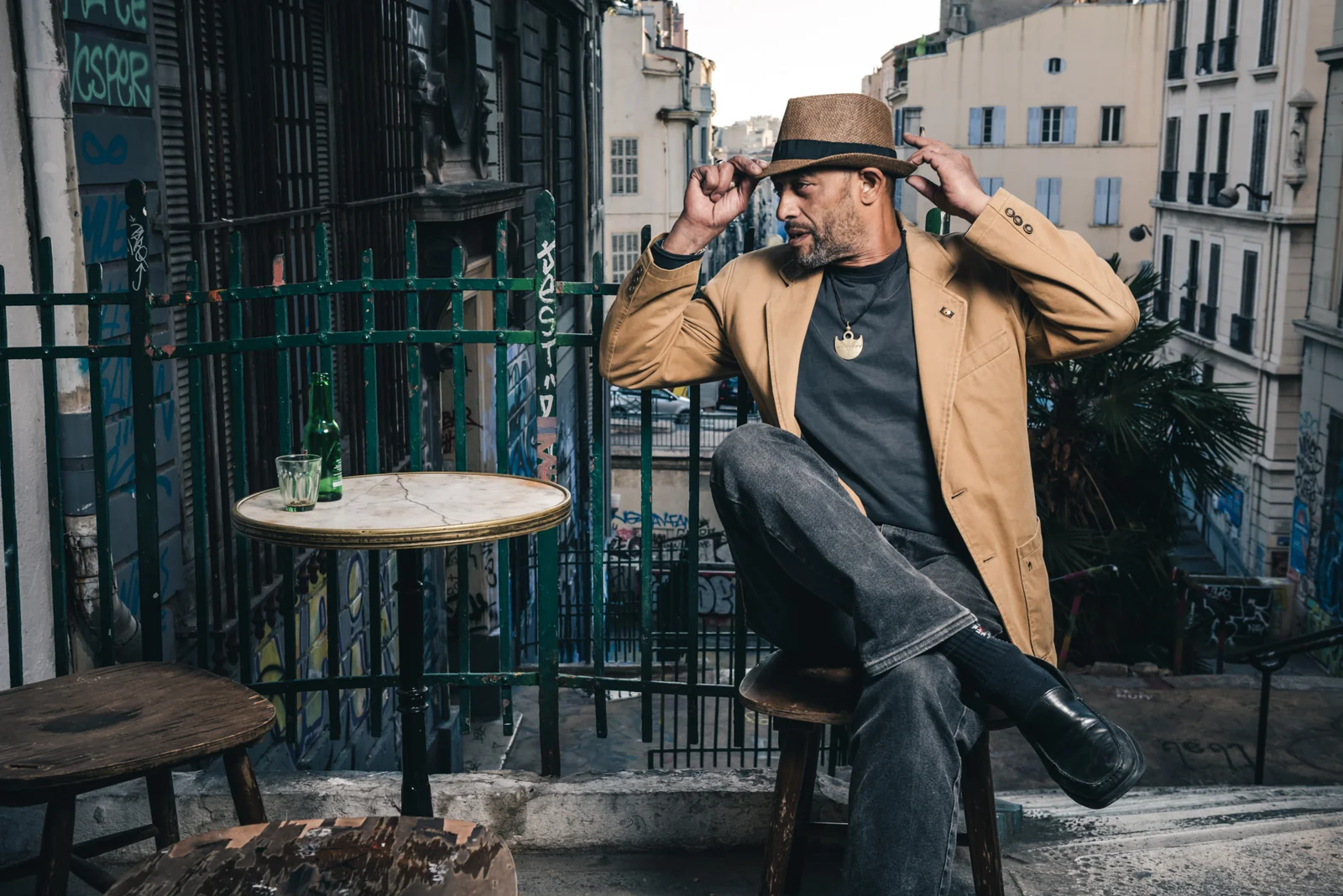 Man in a tan jacket raising his hands beside his hat at an outdoor café.