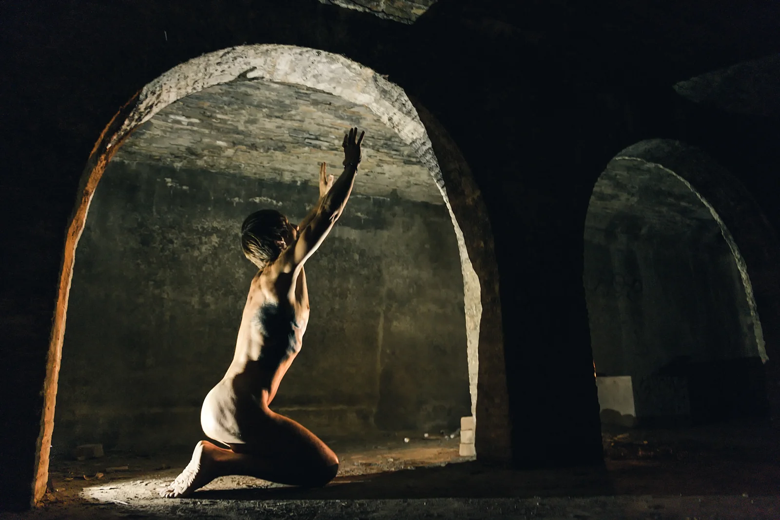 Fine-art nude figure kneeling with one arm raised in a vaulted room.