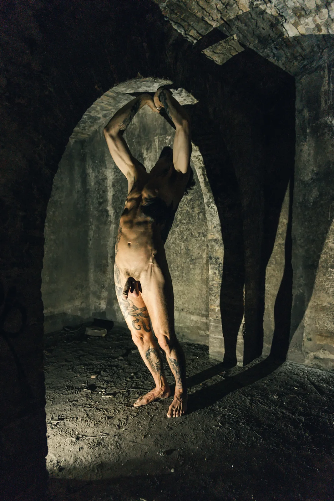 Fine-art nude figure reaching overhead in a dark stone doorway.