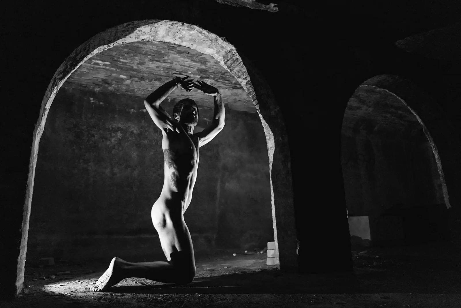 Black-and-white nude figure kneeling with arms curved overhead.