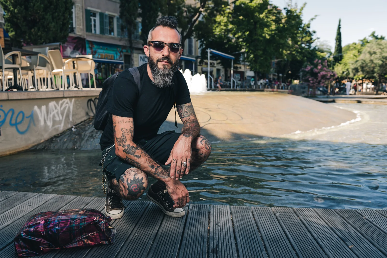 Tattooed man in sunglasses crouching beside a city fountain.