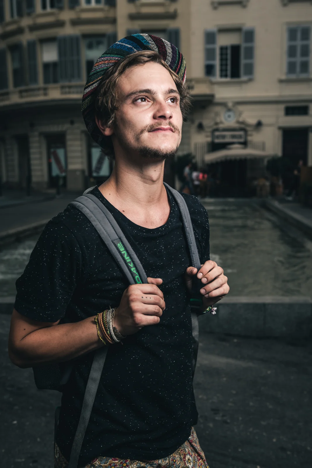 Young man holding backpack straps on a shaded street.
