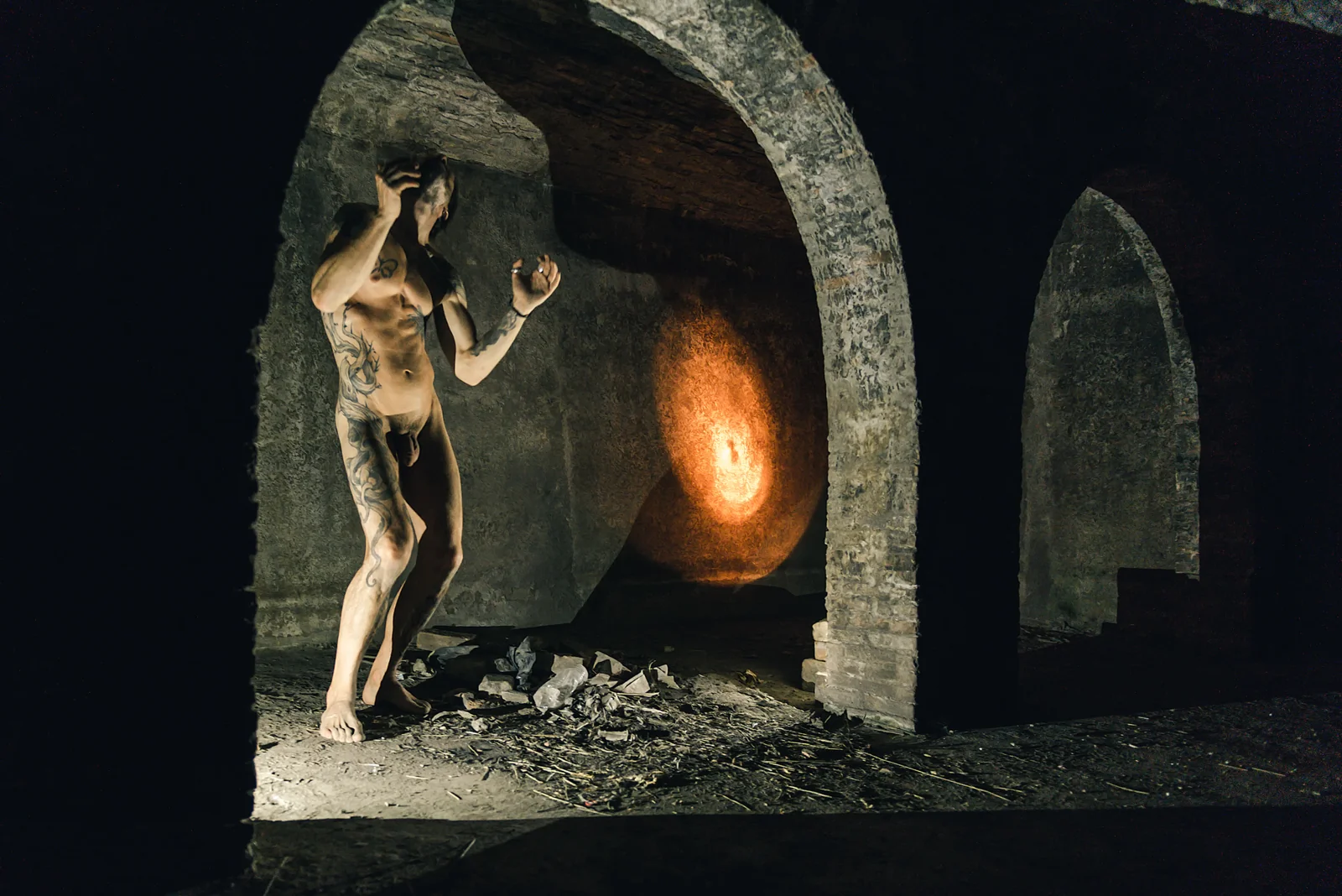 Fine-art nude figure standing in a twisting pose beside warm wall light.