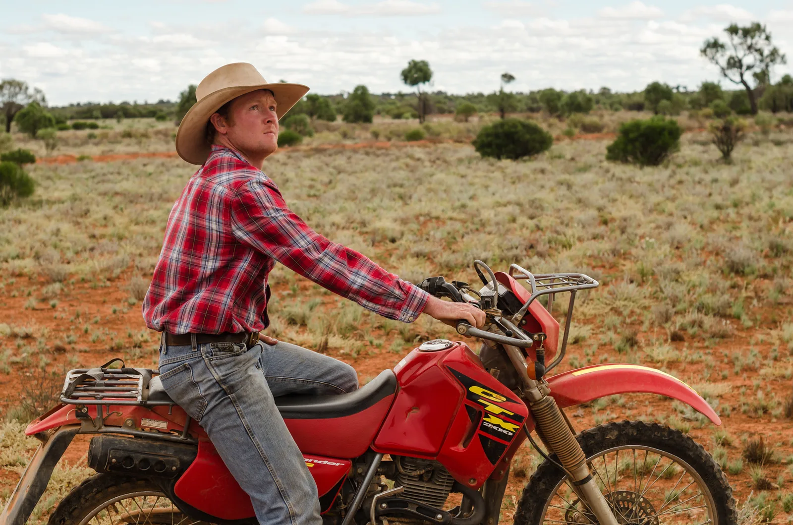 Stockman in a red checked shirt riding a red dirt bike.