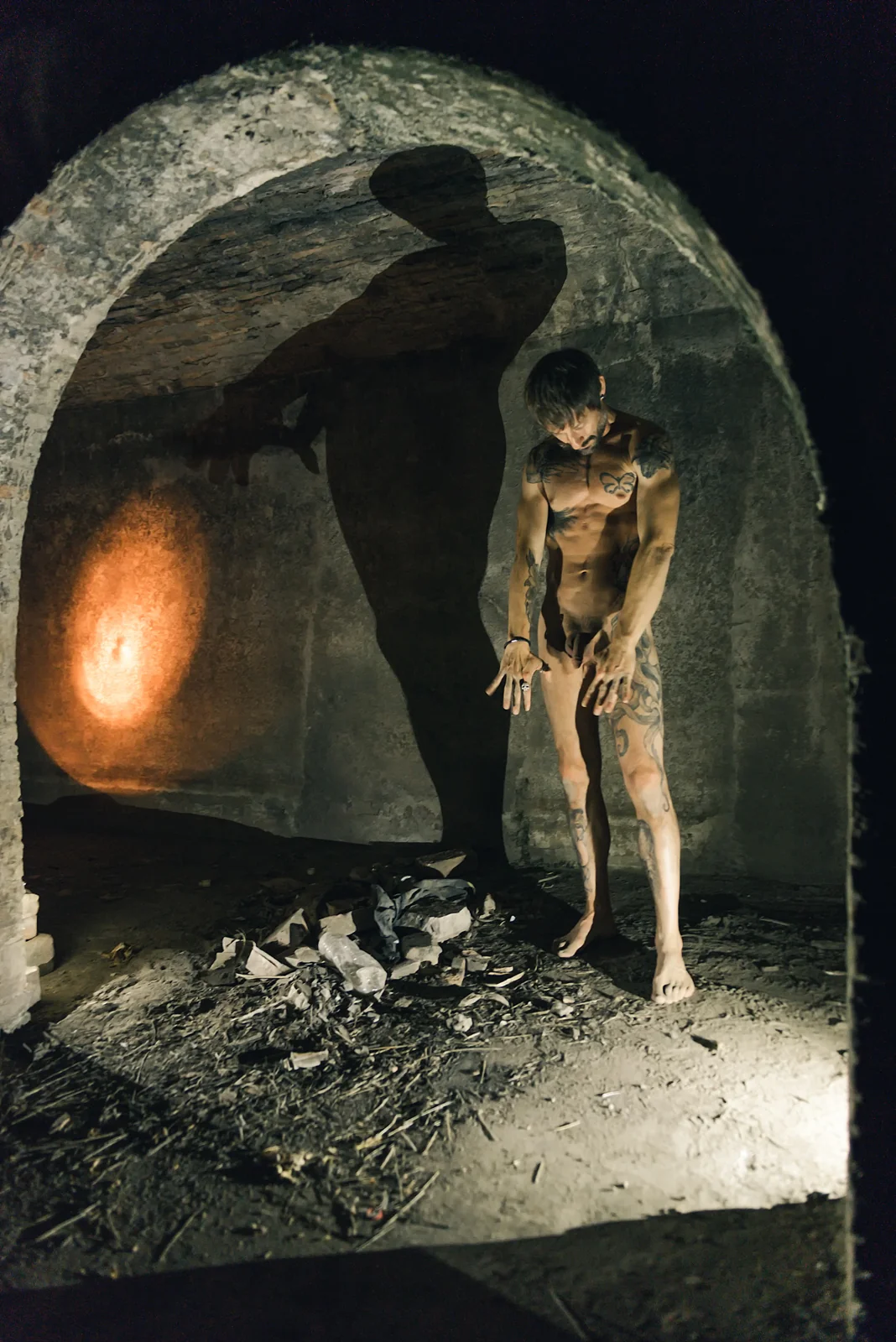 Fine-art nude figure bowing beneath a stone arch with a tall wall shadow.