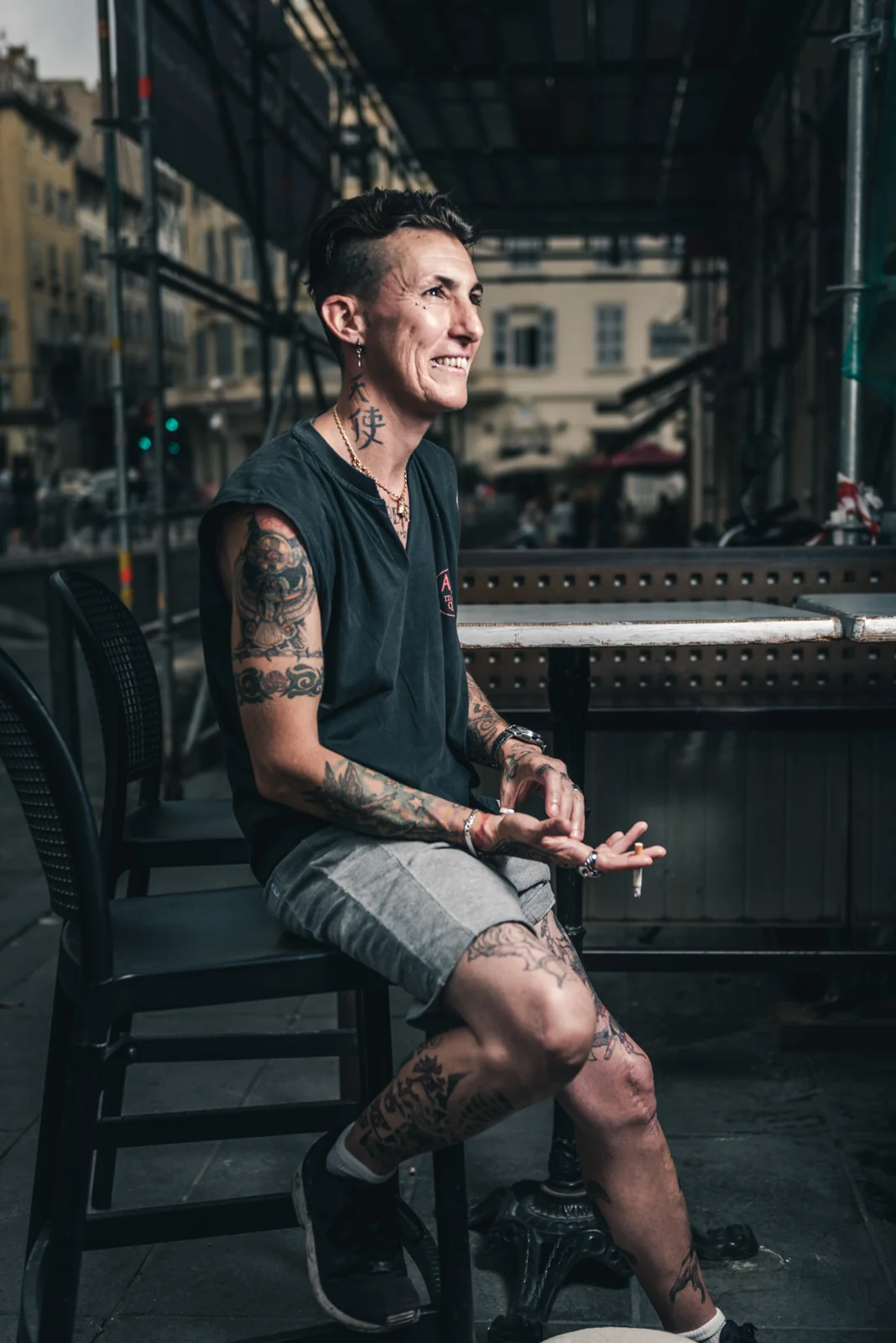 Tattooed man seated on a stool outside a building.