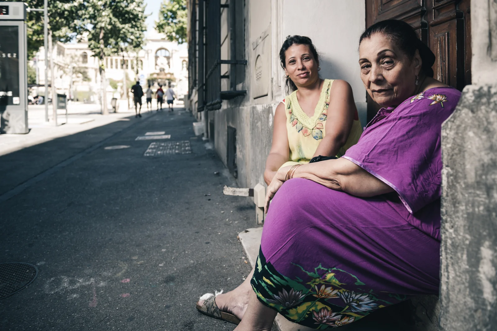 Two women seated beside a building on a quiet sidewalk.
